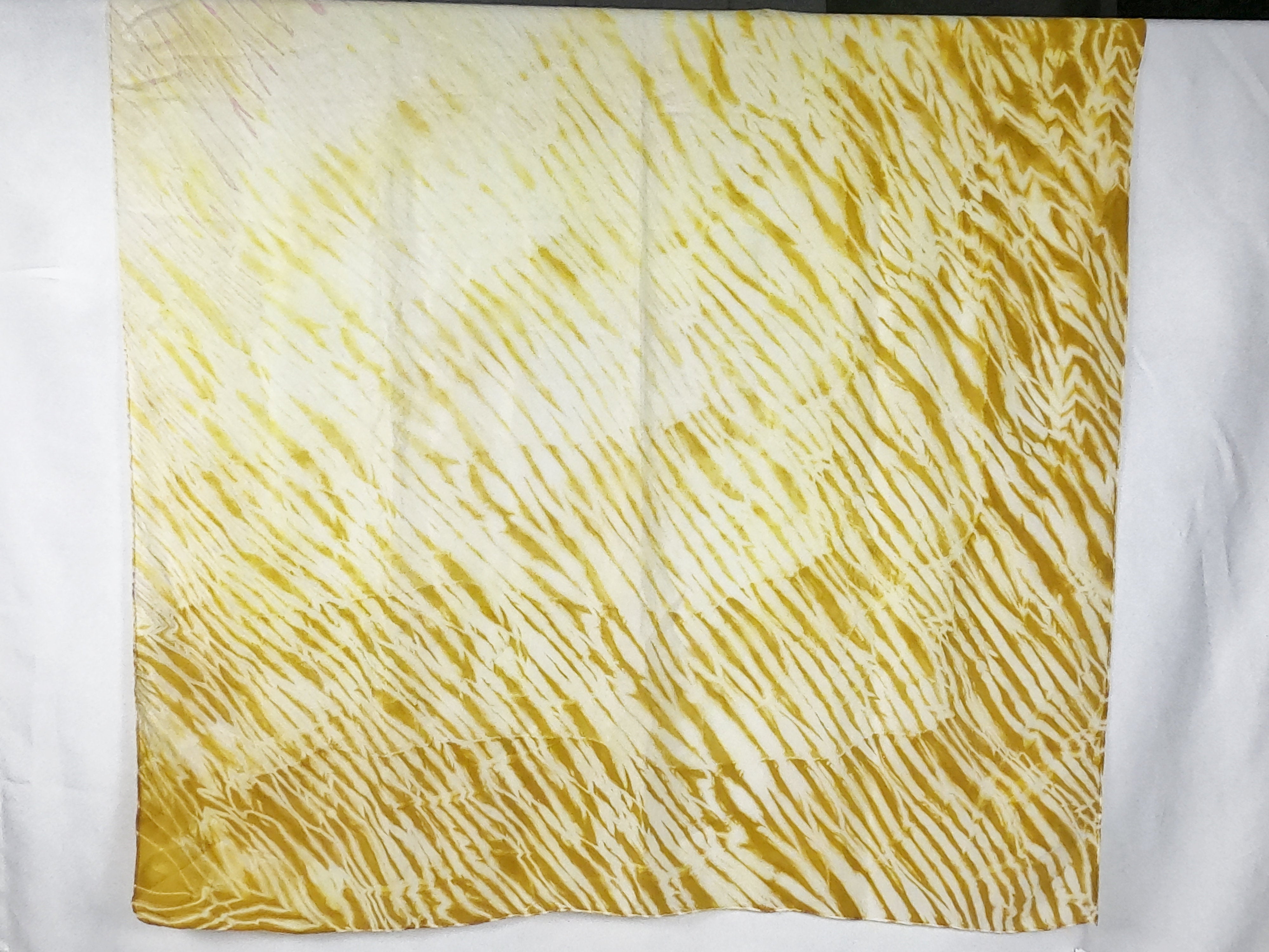 A silk square shawl laid flat. It is dark yellow in colour with a ripple like print, gradually fading from dark to light in an ombre 